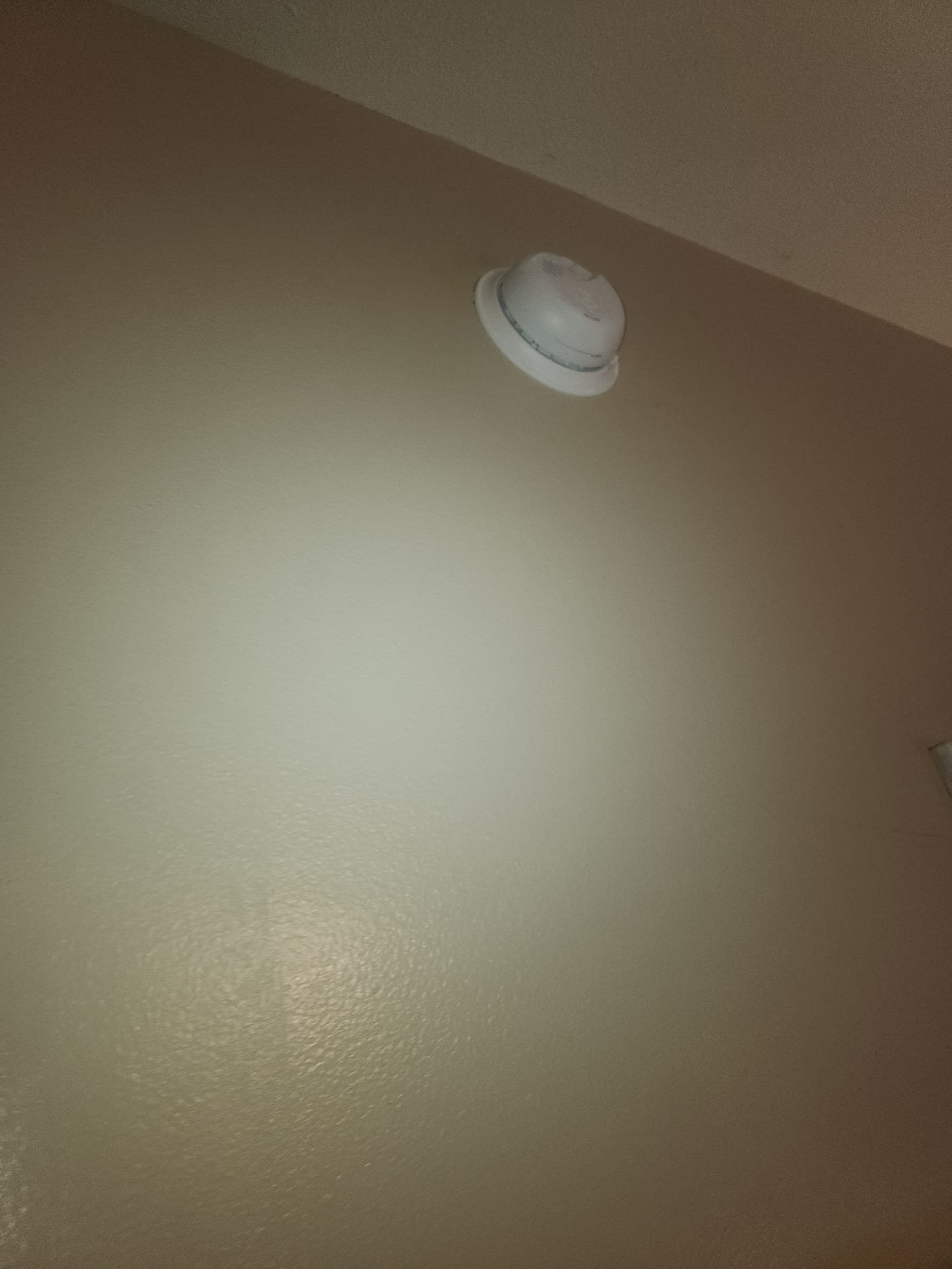 A worn smoke detector mounted near the ceiling before maintenance. It has visible signs of aging and discoloration at the edges.
