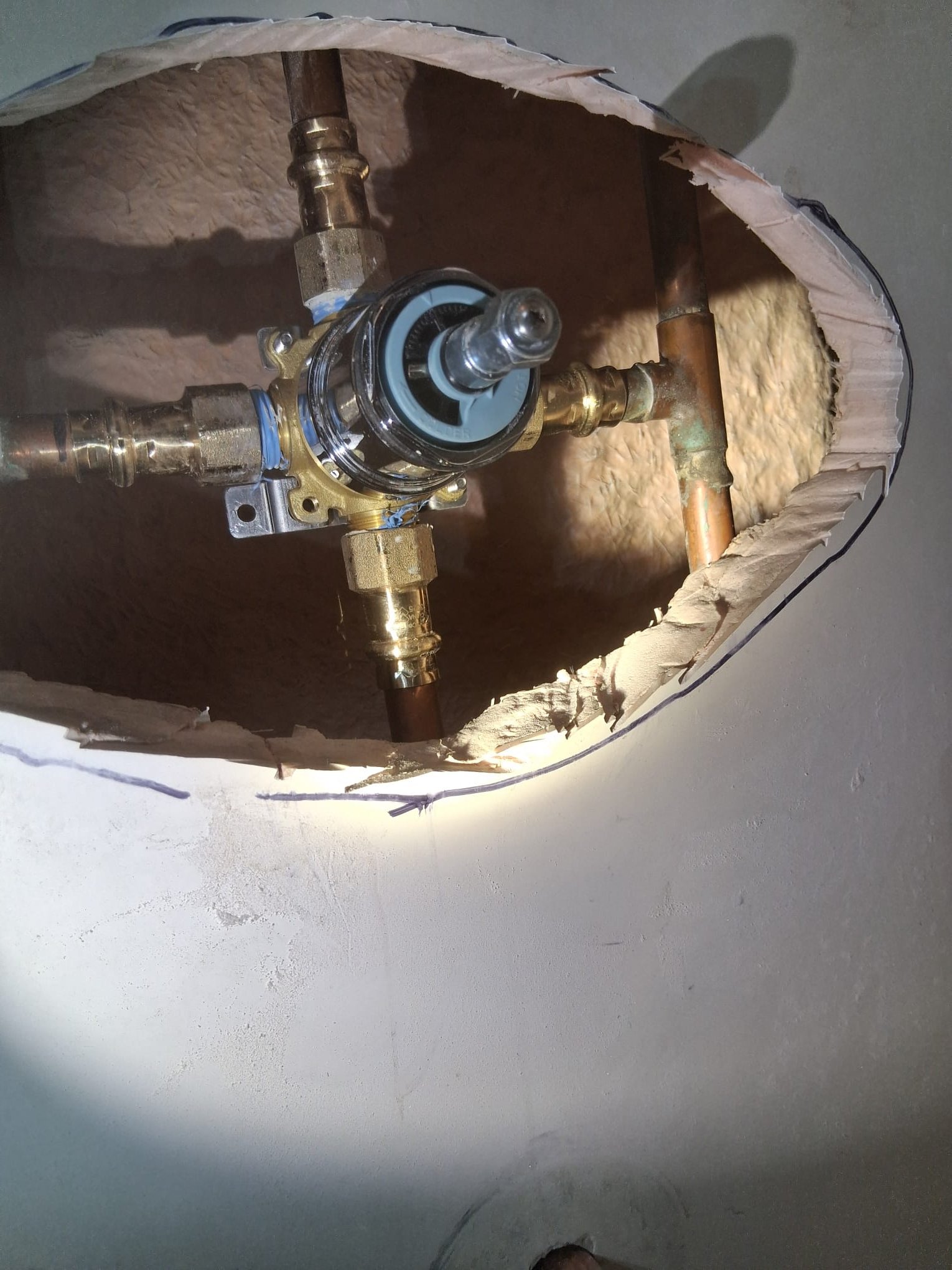 This image shows a plumbing installation in progress, specifically the rough-in stage for a shower or bathtub valve. The wall has been cut open to expose the plumbing work. The visible components include copper pipes and a newly installed brass mixing valve (shower valve), which appears to be in excellent, unused condition.