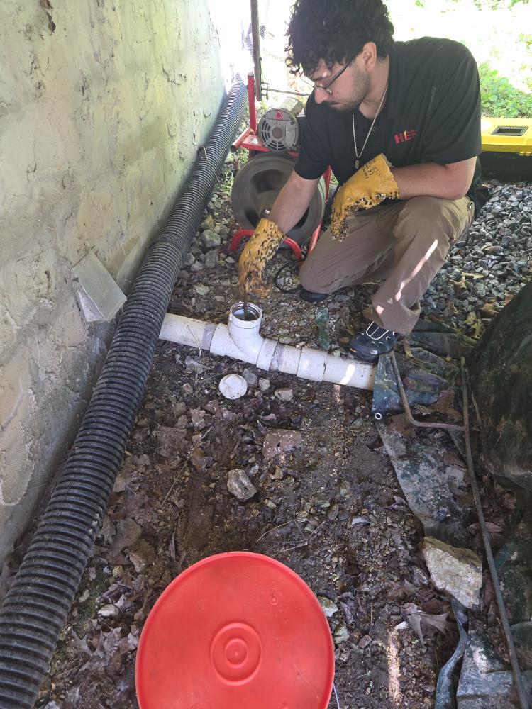 HEP technician working on outdoor drain cleanout near house foundation