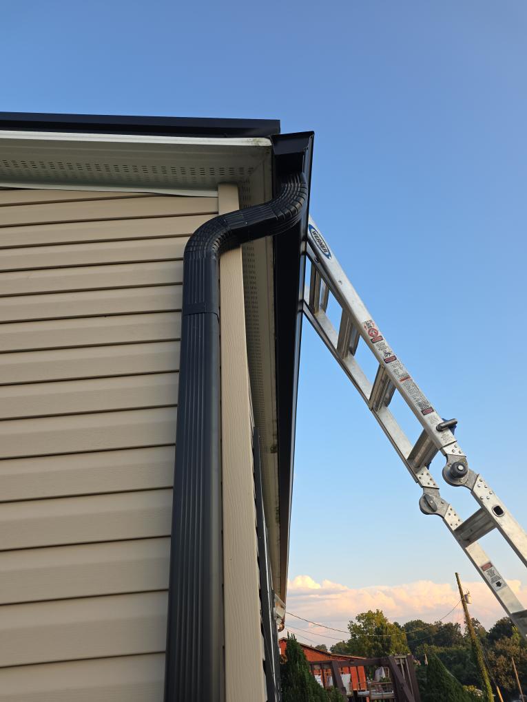 Corner of a house with a newly installed black gutter and downspout, with a ladder still in place